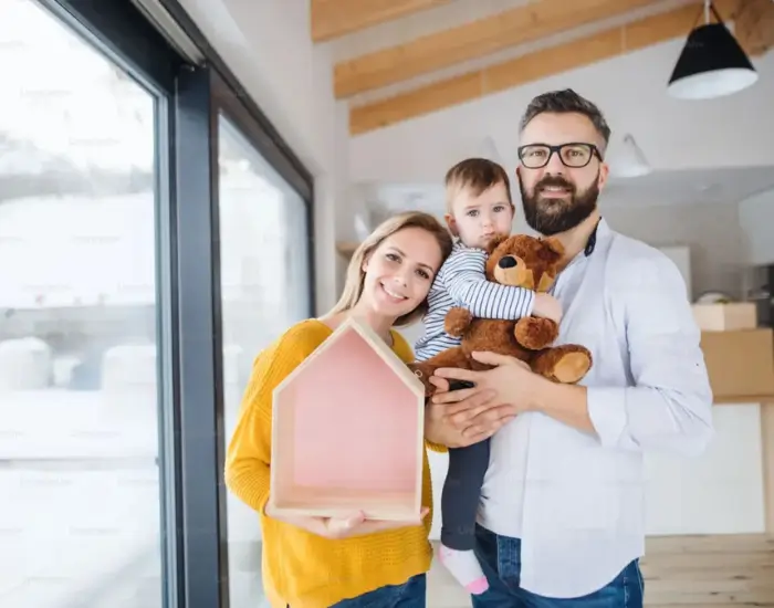 A smiling family, with a man holding a child and a woman holding a small pink house model.
